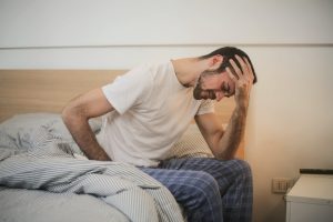 A man holding his forehead while sitting on the edge of his bed.