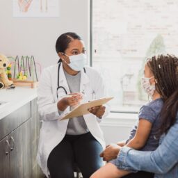 With a protective mask on, a female pediatrician talks to a young patient's mother about the woman's daughter's medical conditions. They are wearing protective masks during an office visit.