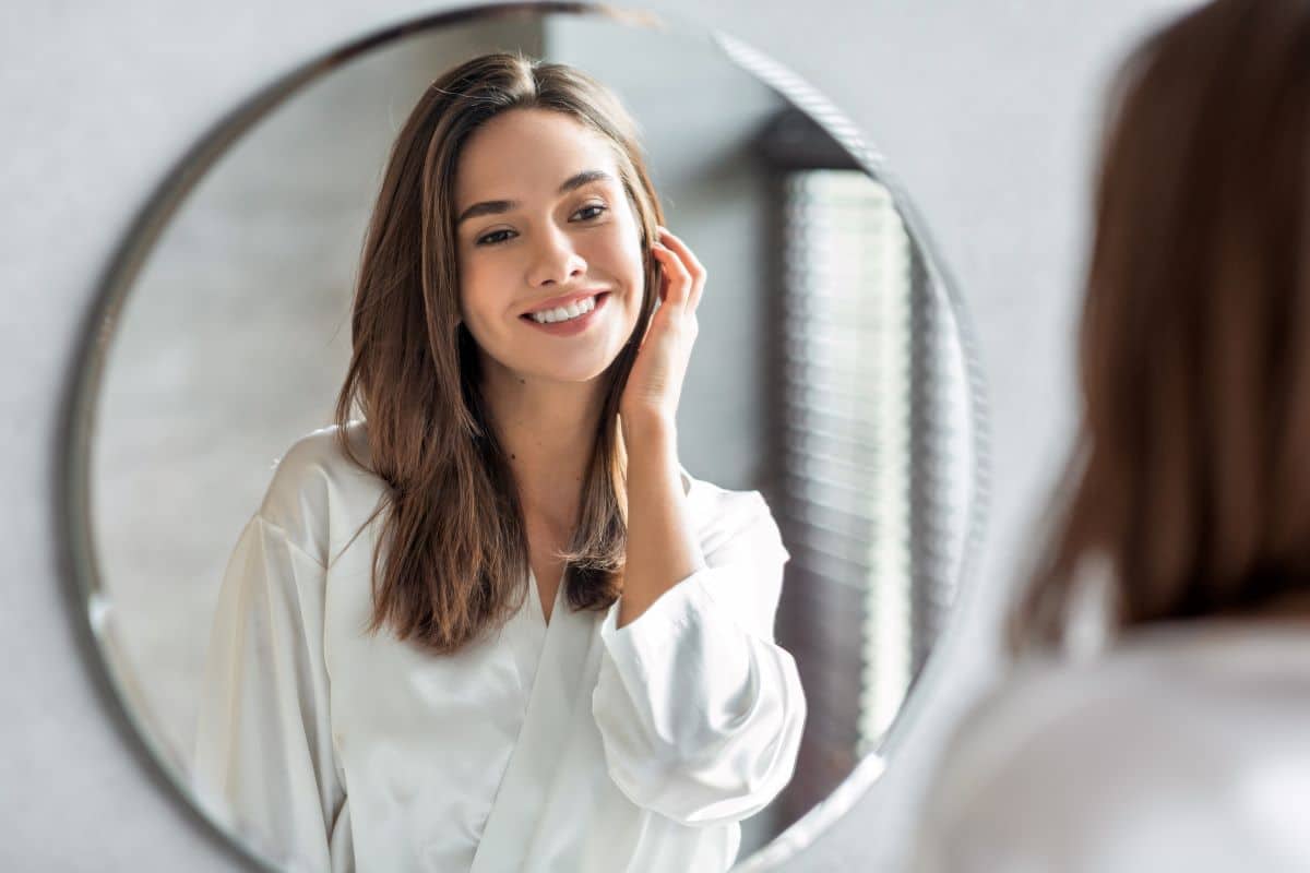 Woman with brunette hair and eyes looking into a bathroom mirror and smiling at herself. One hand is up against her cheek, lightly touching her long hair. The other hand is at her side and out of frame.  