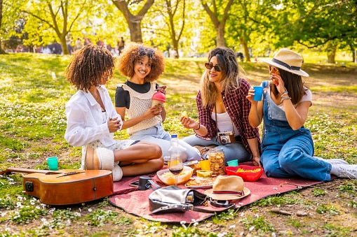 Four young women enjoying a sunny day picnic in the park, laughing, chatting, eating delicious food, and sipping refreshing drinks together.